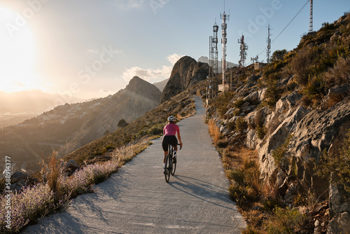 Fit female cyclist wearing cycling kit and helmet riding on the road on a gravel bike at sunset.Empty mountain road. Sports motivation image.Calpe town in Spain.
