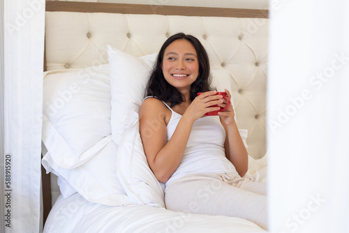 Happy asian woman sitting up in bed holding tea and smiling in bedroom in the morning