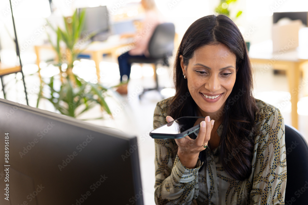 Happy biracial casual businesswoman at table talking on smartphone in modern office Happy biracial casual businesswoman at table talking on smartphone in modern office
