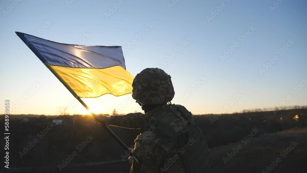Ukrainian army man stands at the peak of hill waving blue-yellow banner against sunset. Male soldier in camouflage uniform lifting flag of Ukraine as a symbol of victory against russian aggression