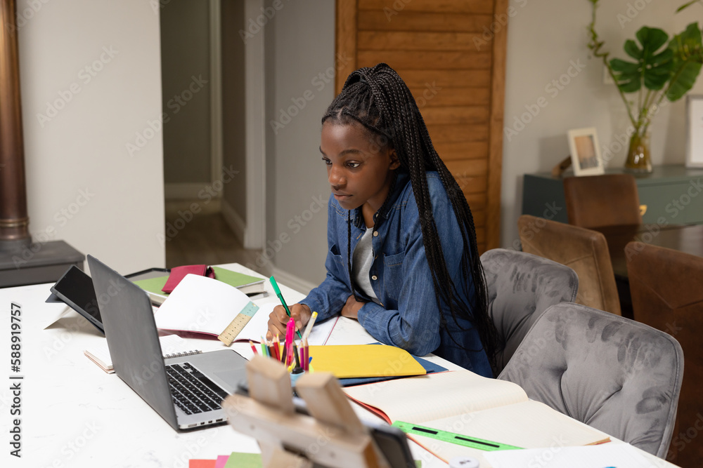 Happy african american teenager girl sitting by table, doing homework and using laptop Happy african american teenager girl sitting by table, doing homework and using laptop