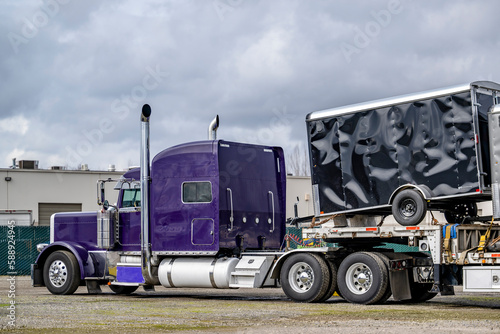 Violet classic big rig American bonnet semi truck with loaded step down semi trailer standing on the industrial warehouse parking lot waiting for the next freight start