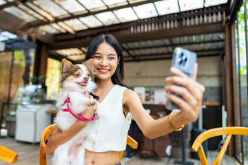 Young Asian woman using mobile phone taking selfie with her chihuahua dog at pets friendly cafe. Domestic dog with owner have fun urban outdoor lifestyle on summer vacation. Pet Humanization concept.