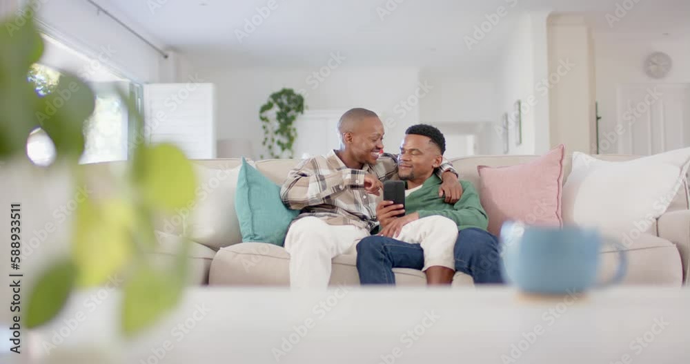 Happy african american gay male couple sitting on sofa, embracing and using smartphone, slow motion Happy african american gay male couple sitting on sofa, embracing and using smartphone, slow motion