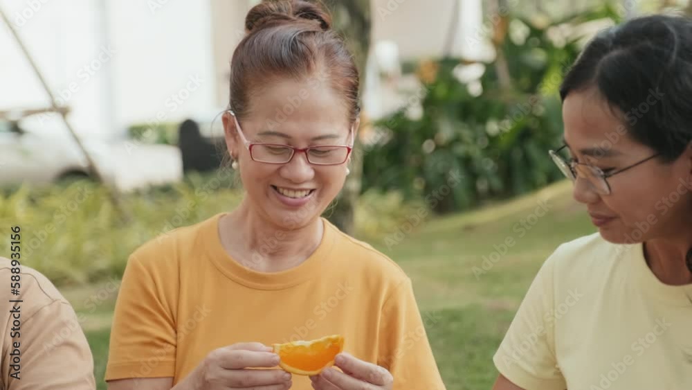 Tilt up shot of Asian woman sharing orange slices with family members while enjoying fruit on summer picnic in park Tilt up shot of Asian woman sharing orange slices with family members while enjoying fruit on summer picnic in park