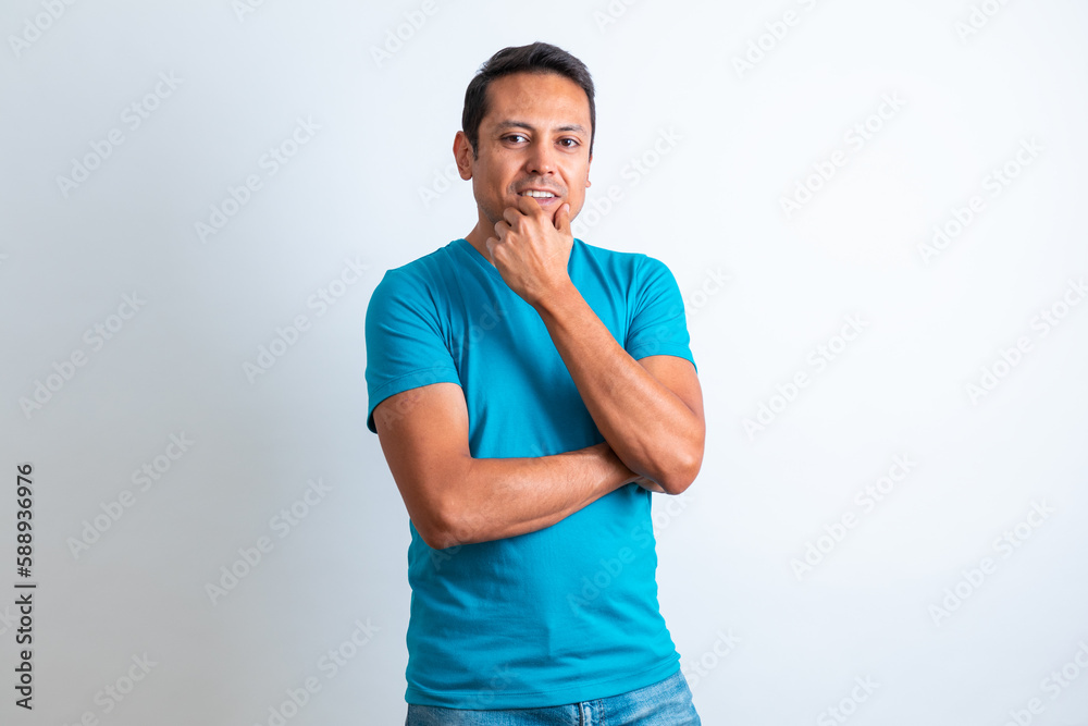 Hombre joven moreno, con ropa casual playera azul, sonriendo y tocando la barbilla con fondo blanco Hombre joven moreno, con ropa casual playera azul, sonriendo y tocando la barbilla con fondo blanco