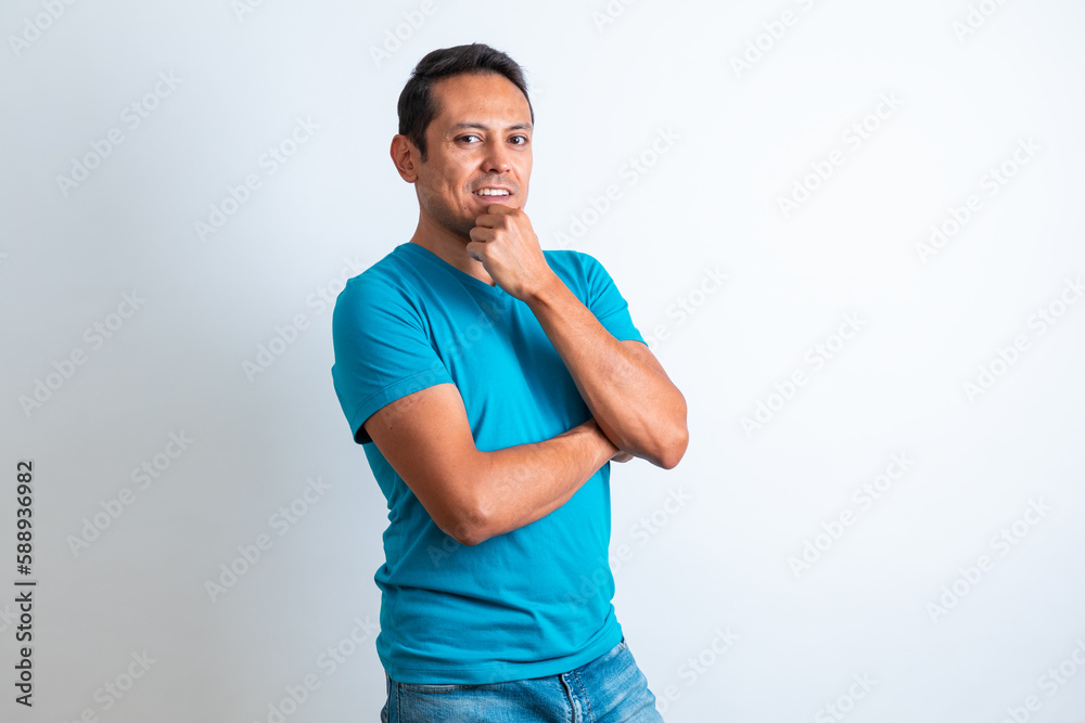 Hombre joven moreno, con ropa casual playera azul, sonriendo y tocando la barbilla con fondo blanco Hombre joven moreno, con ropa casual playera azul, sonriendo y tocando la barbilla con fondo blanco