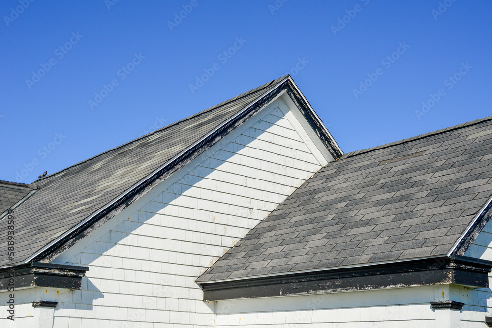 Multiple peaked and layered roofs on a white vintage wooden house. The eave is painted black. The shingles on the roof are grey in color. The old building has wood horizontal clapboard siding.