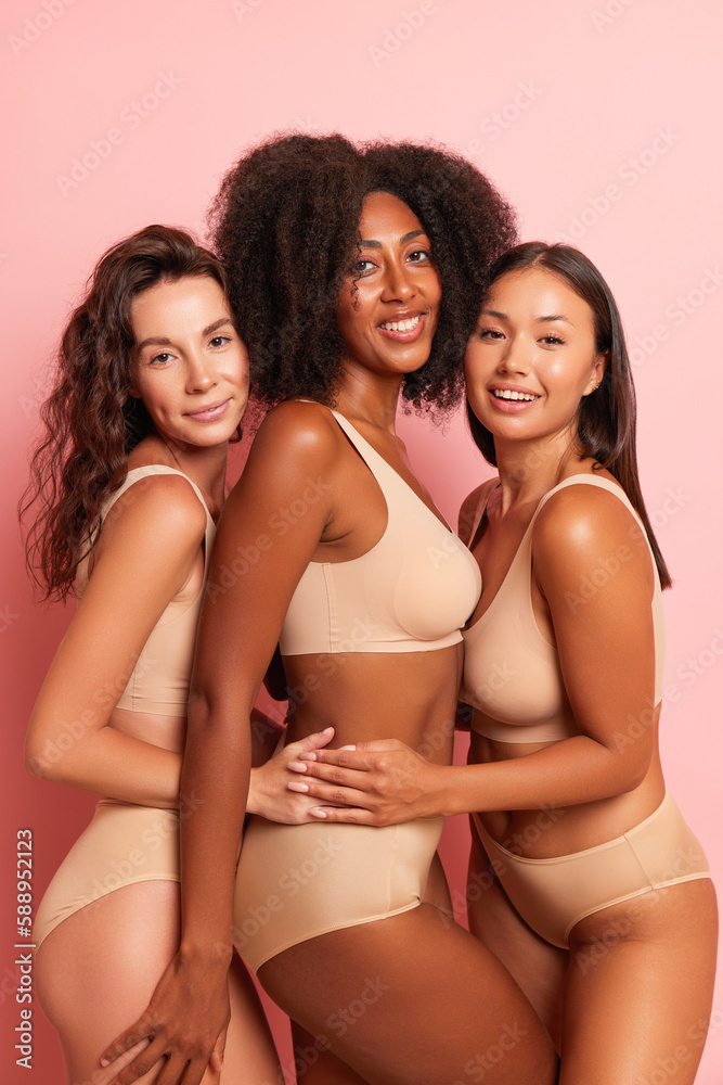 Three lovely girls dressed in nude-colored underwear posing on pink background, African-American in middle, Asian and European models are at her sides, girls having fun, happy time concept, high