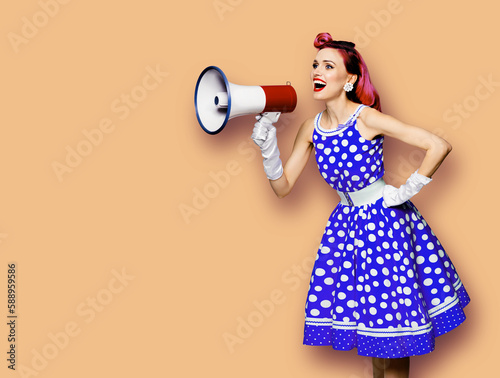 Purple haired woman holding red megaphone, shout advertise something. Girl in blue pin up style with mega phone loudspeaker. Latte beige background with mock up. Female model in retro fashion dress.