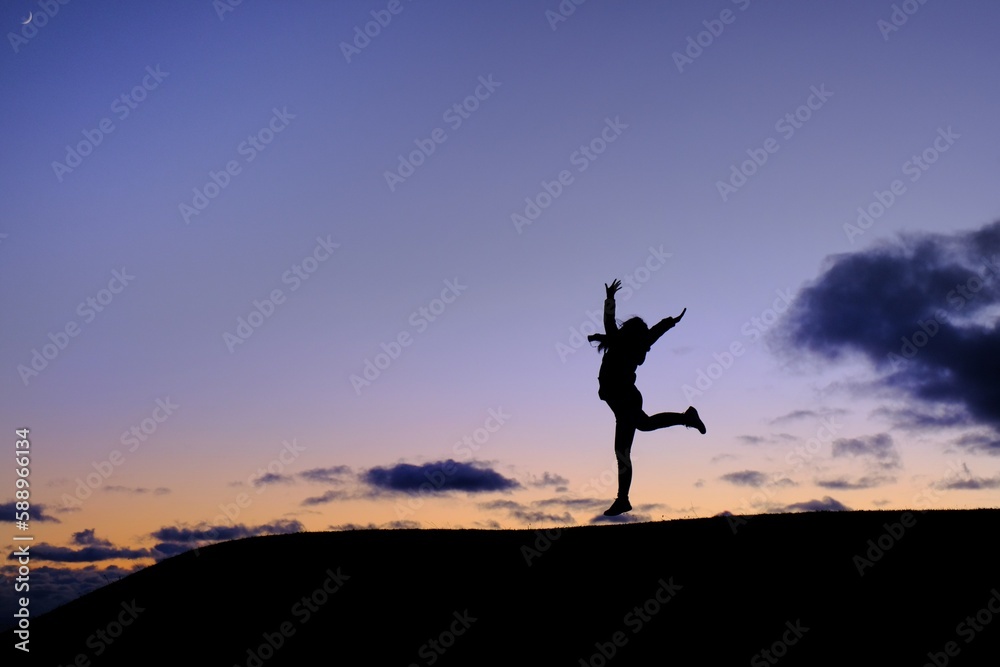 A silhouette of a woman in gym uniform jumping on top of a hill in celebration after completing her exercise with blue and orange sky in the background with a crescent moon at the top left corner.