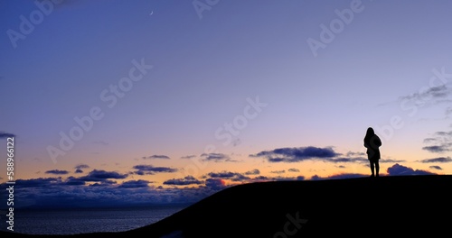 A silhouette of a woman standing alone one top of a hill by the sea, watching sunset with cloudy colorful sky, feeling peaceful and calm.