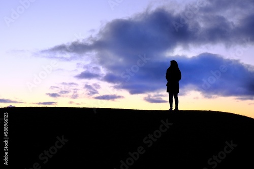 A silhouette of a woman standing alone one top of a hill by the sea, watching sunset with cloudy colorful sky, feeling peaceful and calm.