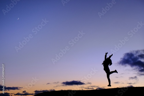 A silhouette of a woman in gym uniform jumping on top of a hill in celebration after completing her exercise with blue and orange sky in the background with a crescent moon at the top left corner. © Sirichai