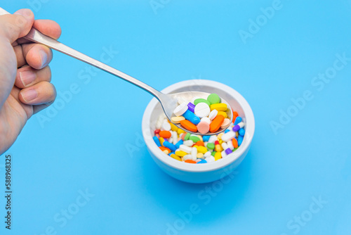 A mortar is placed on a blue background and filled with colorful pills capsules, a spoon and an empty space to use as a copyspace. human hand is floating with a spoon.