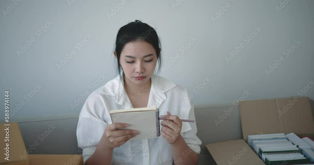 Handheld shot, Asian young woman sits and checking books in box and write on notebook after move into new home Handheld shot, Asian young woman sits and checking books in box and write on notebook after move into new home