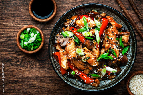 Stir fry chicken with paprika, mushrooms, green chives and sesame seeds in ceramic bowl. Asian cuisine dish. Wooden kitchen table background, top view