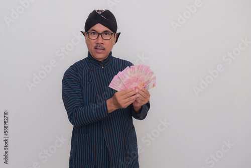 A middle-aged man wearing Javanese traditional outfit holds some money; serious, happy expression. photo
