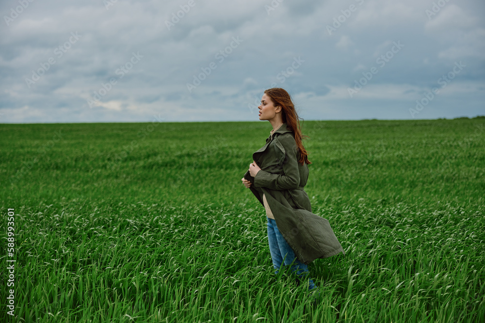 Fototapeta premium a red-haired woman stands in a green field in rainy, cold weather, holding a raincoat in the wind