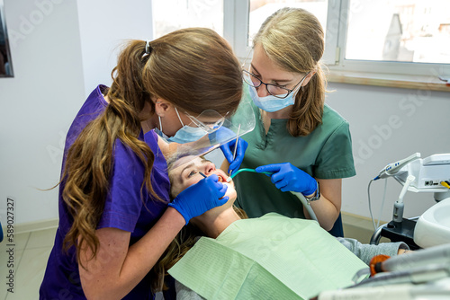 Dentist and assistant in masks and uniforms making professional teeth cleaning female patient