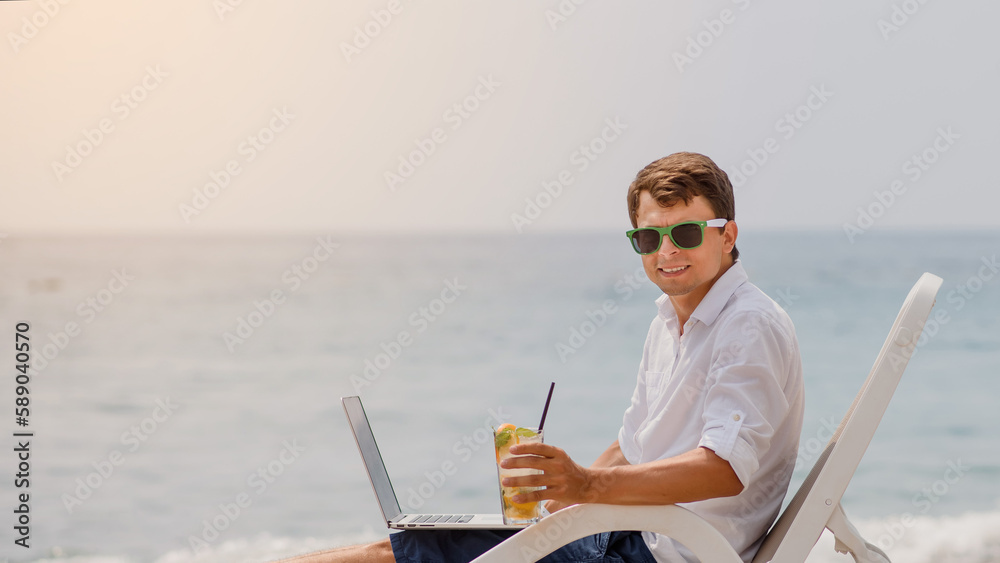 Young man in sunglasses and white shirt is lying on a beach chair on the seaside holding a cocktail and a laptop.