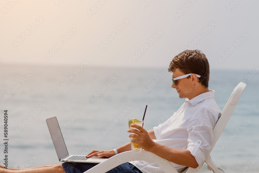 Young man in sunglasses and white shirt is lying on a beach chair on the seaside holding a cocktail and a laptop.