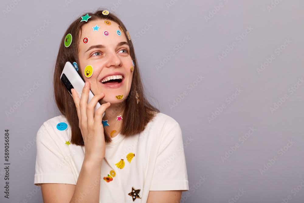 Φωτογραφία Image of smiling positive woman covered with funny stickers posing isolated over gray background, talking on smart phone and laughing, copy space for advertisement