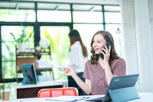 Young business woman on the phone at the office.Business woman texting on the phone and working on laptop.Beautiful young business woman sitting in office.Business woman smiling.