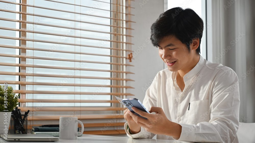 Smiling millennial asian man sitting in home office checking social media, chatting online on smartphone Smiling millennial asian man sitting in home office checking social media, chatting online on smartphone