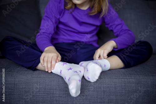 A cute little blonde girl puts white socks with stars on her feet. Independence of children. The development of dress skills in younger schoolchildren