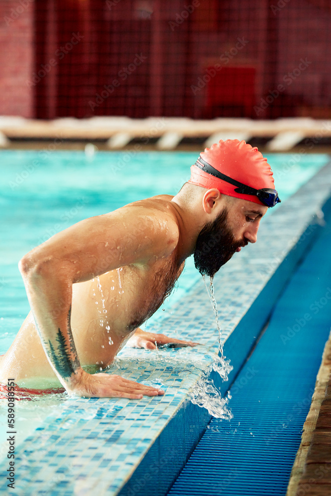 A swimmer in a hat and goggles on the side of the pool, a male swimmer emerges from the pool in a red cap for swimming, flying out of the pool emerges from the swimming training break