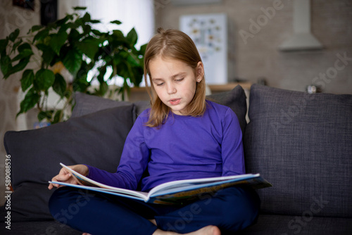 A cute little blonde girl is reading a large colorful book sitting on the sofa in the living room. Self-education of younger schoolchildren  reading literature at home  interesting leisure