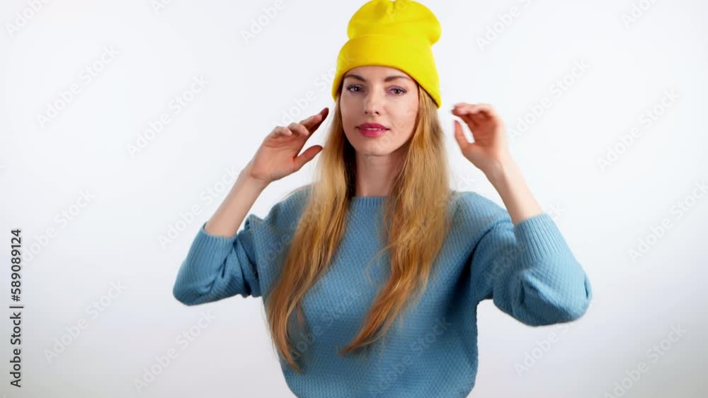 Cheerful young woman dancing. Funny smiling overjoyed blonde lady waving hands in yellow beanie and blue sweater. Indoor studio, emotional 20s girl enjoys dancing isolated on white background. Cheerful young woman dancing. Funny smiling overjoyed blonde lady waving hands in yellow beanie and blue sweater. Indoor studio, emotional 20s girl enjoys dancing isolated on white background.