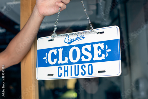 A person hanging up a sign on a restaurant door, reading Close. Closed, dual language, English and Italian. photo