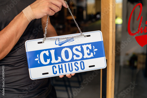 A person hanging up a sign on a restaurant door, reading Close. Closed, dual language, English and Italian. photo
