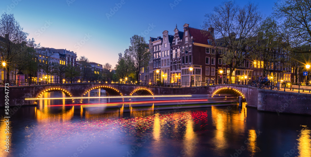 A cityscape of Amsterdam in the Netherlands at dusk, illuminated by light trails reflecting off its river: boats travel under a bridge and buildings line either side.