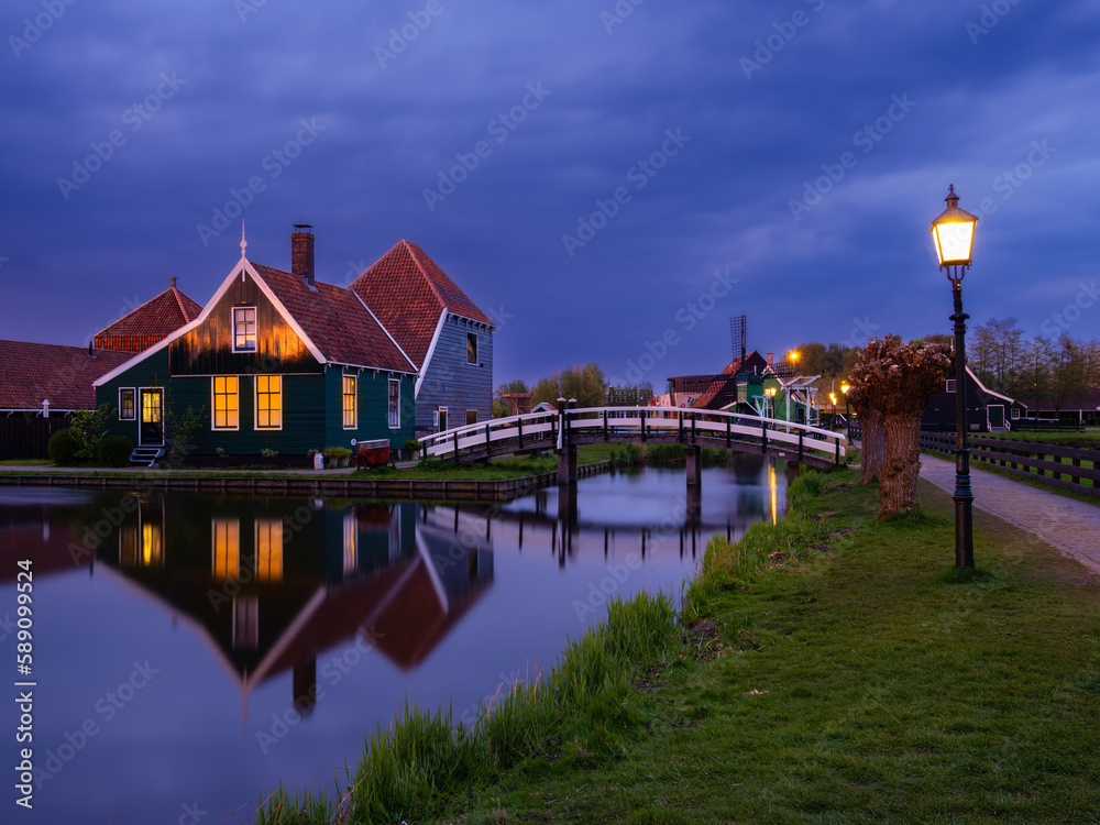 Zaanse Schans, Netherlands - April 25, 2022: At dusk, the illuminated buildings of Zaanse Schans in Holland reflect off the waterway and bridge, creating a beautiful night scene.