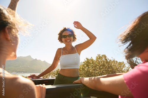 Female Friends Standing Up Through Sun Roof Car And Dancing On Road Trip Through Countryside