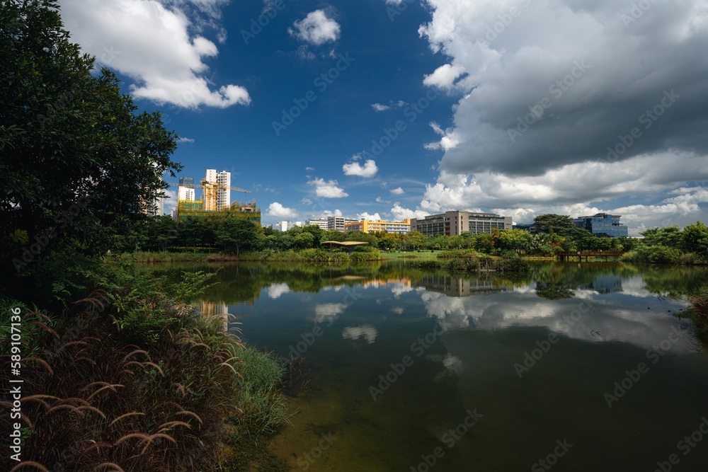 Obraz premium Sky clouds with reflection in a lake surrounded by trees and buildings in the background