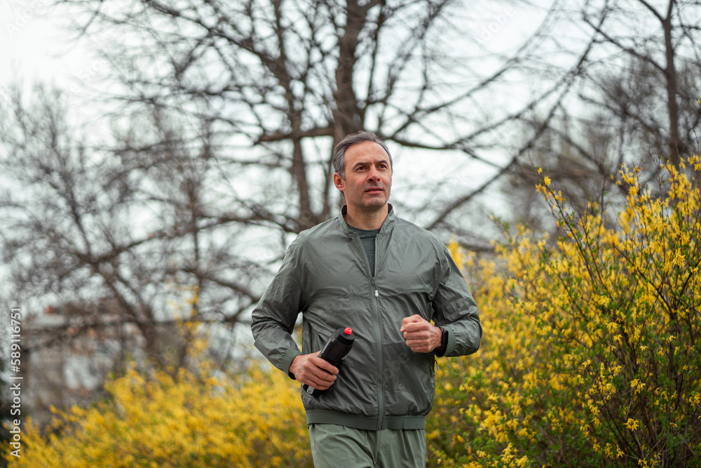 Obraz premium A middle shot of a fit middle-aged man jogging in a city park with a yellow flower bush in the background.