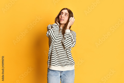 Attractive young girl in striped sweater listening to music in heaphones and looking away with dreamy look against yellow studio background Concept of youth, emotions, facial expression, lifestyle
