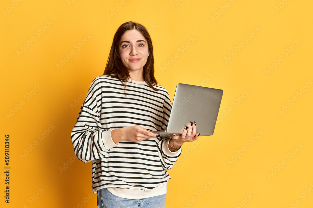 Young smiling girl in striped sweater posing with laptop against yellow studio background. Job fair and student life. Concept of youth, emotions, facial expression, lifestyle, education, business Young smiling girl in striped sweater posing with laptop against yellow studio background. Job fair and student life. Concept of youth, emotions, facial expression, lifestyle, education, business
