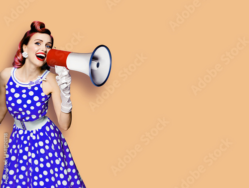 Portrait image of purple haired woman holding mega phone, shout advertise something. Girl in blue pin up style dress with mega phone loudspeaker. Isolated latte beige background. Beauty model.