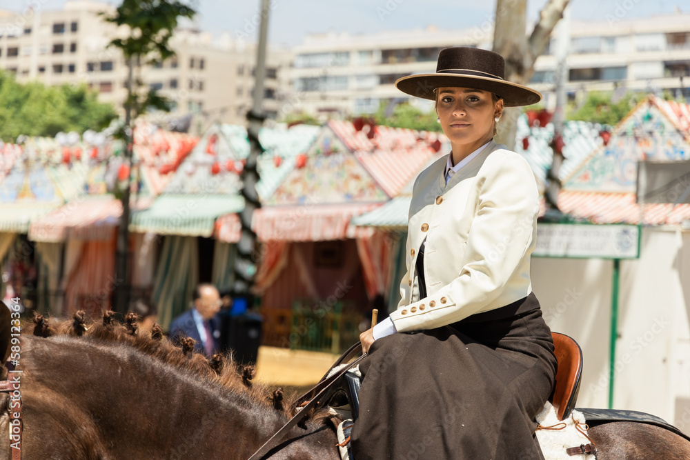 Young woman dressed in traditional flamenco cowgirl costume riding a horse at a fair looking at the camera