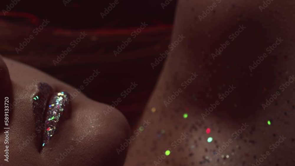 Vertical video. Glitter skin. Makeup art. Nightclub look. Cropped closeup of neon blue red color light woman face with sparkling metallic silver lips on dark.