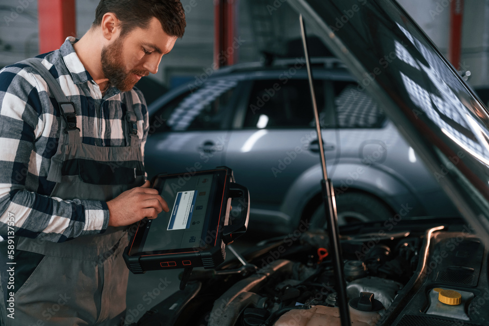 Standing near the car with hood opened. Holding tablet. Man in uniform is working in the auto salon Standing near the car with hood opened. Holding tablet. Man in uniform is working in the auto salon