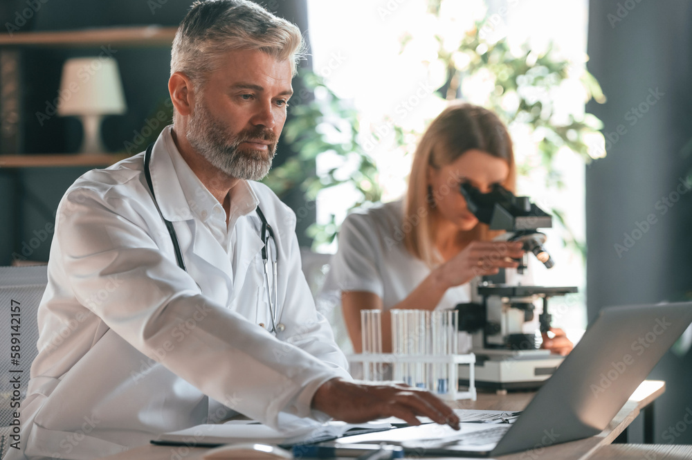 Using microscope. Two clinic workers are indoors together. Senior man with young woman Using microscope. Two clinic workers are indoors together. Senior man with young woman
