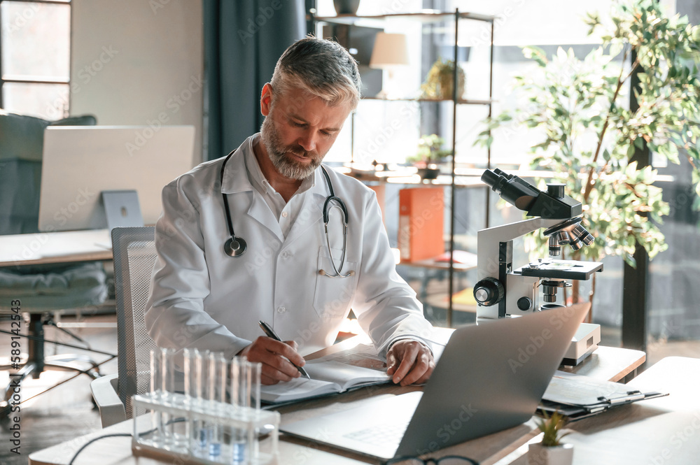Writing information into document. Senior doctor in white coat is at work indoors Writing information into document. Senior doctor in white coat is at work indoors