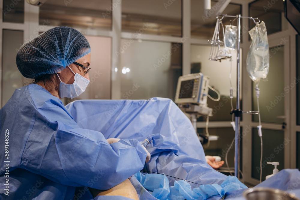 An experienced angiosurgeon operates on the leg of a patient with varicose veins. Doctor in a protective mask and an operating gown concentrates on the operation on the patient's leg. An experienced angiosurgeon operates on the leg of a patient with varicose veins. Doctor in a protective mask and an operating gown concentrates on the operation on the patient's leg.