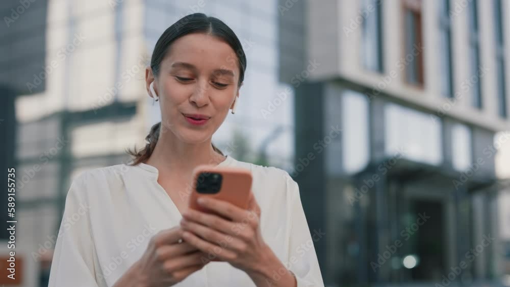 Cheerful Beautiful Woman Chatting on Video Call Using Wireless Earphones at Street Close Up. Happy Young Lady Making Online Conference with Friends at Smartphone. Stylish Smiling Girl Looking Screen Cheerful Beautiful Woman Chatting on Video Call Using Wireless Earphones at Street Close Up. Happy Young Lady Making Online Conference with Friends at Smartphone. Stylish Smiling Girl Looking Screen